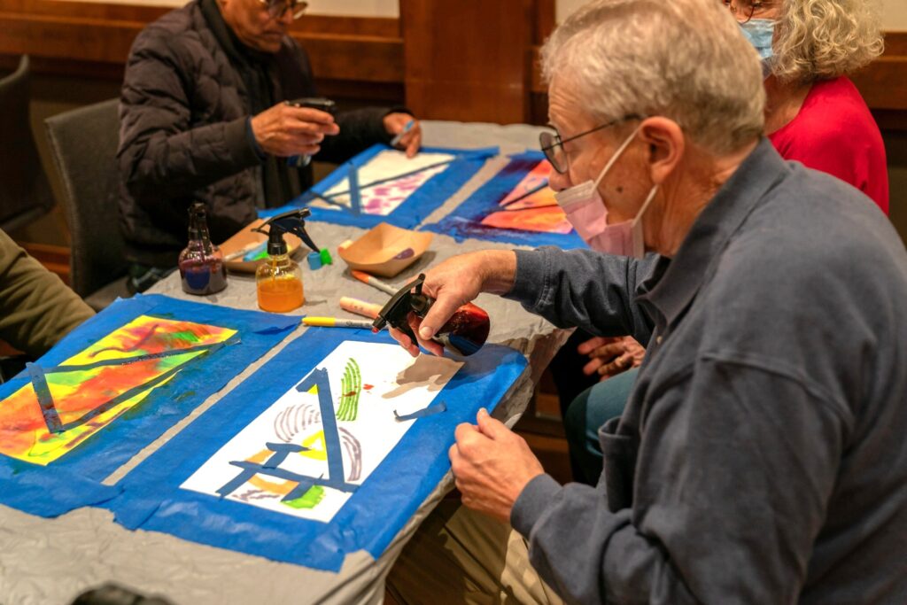 An older adult man sprays his painting while sitting at a table.