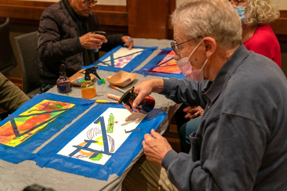 An older man sits at a table with other older adults while spraying liquid on an abstract painting. 