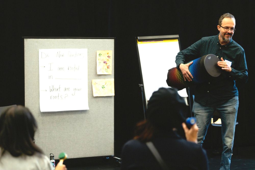 A mididle-aged man with dark short hair stands near a posterboard and holds a drum. People are watching him.