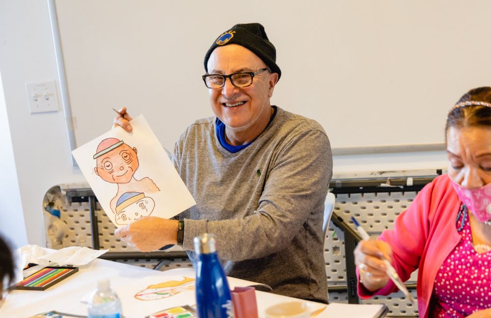 A photo of an older adult white man smiling at the camera and sitting at a table holding a drawing he made. He is wearing a black beanie, glasses, and grey long sleeved shirt.