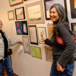 A photo of two older adult Asian/Pacific Islander women standing in front of a wall of artwork. They are smiling and not looking directly at the camera. One woman has long, silver/grey hair and is wearing a grey sweater and jeans. She is carrying a brown purse on her shoulder. The other woman has grey/silver hair pulled back. She is wearing a black shirt with a silver/white coat and jeans. She is carrying a black over the shoulder bag.