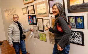 A photo of two older adult Asian/Pacific Islander women standing in front of a wall of artwork. They are smiling and not looking directly at the camera. One woman has long, silver/grey hair and is wearing a grey sweater and jeans. She is carrying a brown purse on her shoulder. The other woman has grey/silver hair pulled back. She is wearing a black shirt with a silver/white coat and jeans. She is carrying a black over the shoulder bag.