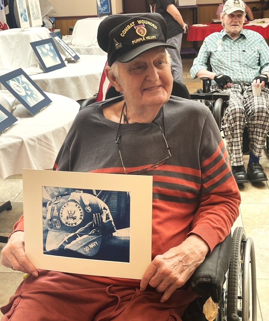 A photo of an older adult veteran white man sitting in his wheelchair and holding a photography work of a hat. The man is wearing a veterans hat and a grey and orange striped shirt. He is smiling and not looking directly at the camera.