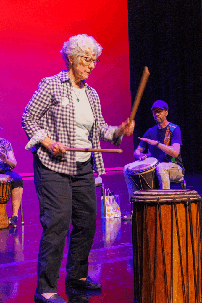 A close-up of an older woman banging on drums with sticks.