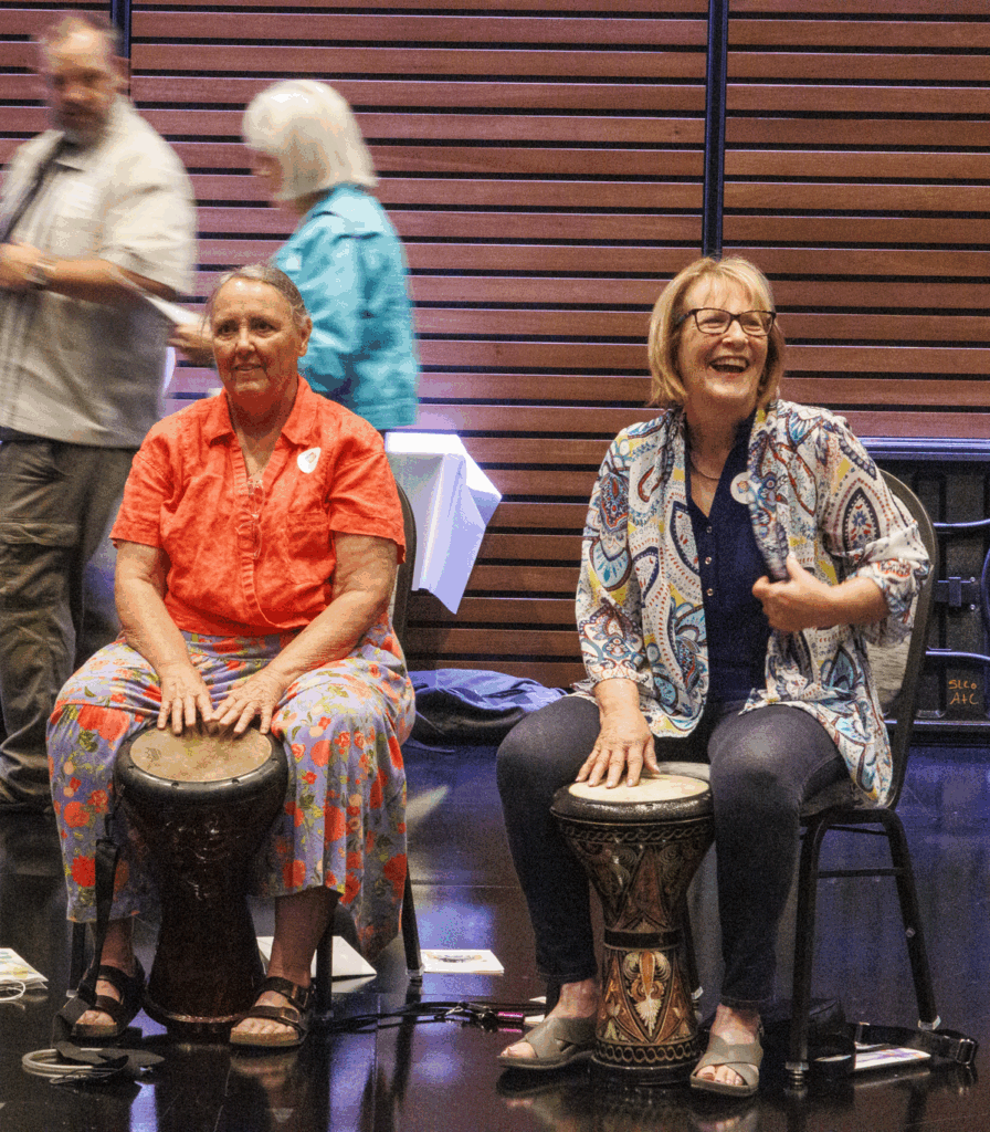 Two older adult women sit on stools and drum.