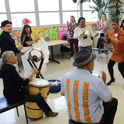 A group of older adults dance and play drums in a large room with lots of windows.