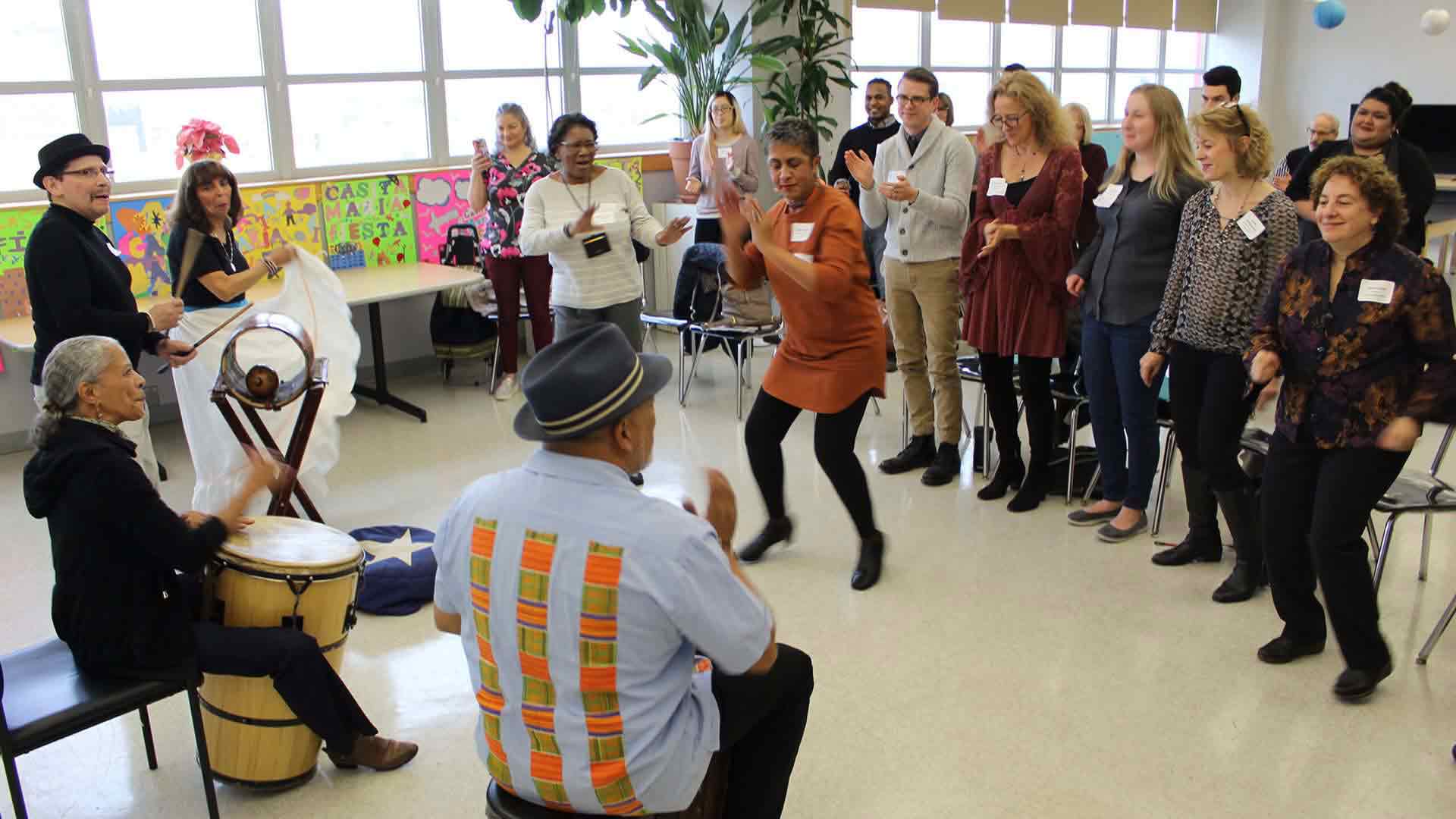 A group of older adults dance and play drums in a large room with lots of windows.