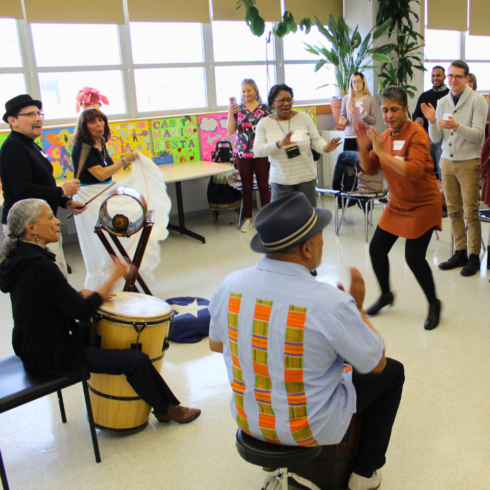 A group of older adults dance and play drums in a large room with lots of windows, plants, and artwork.