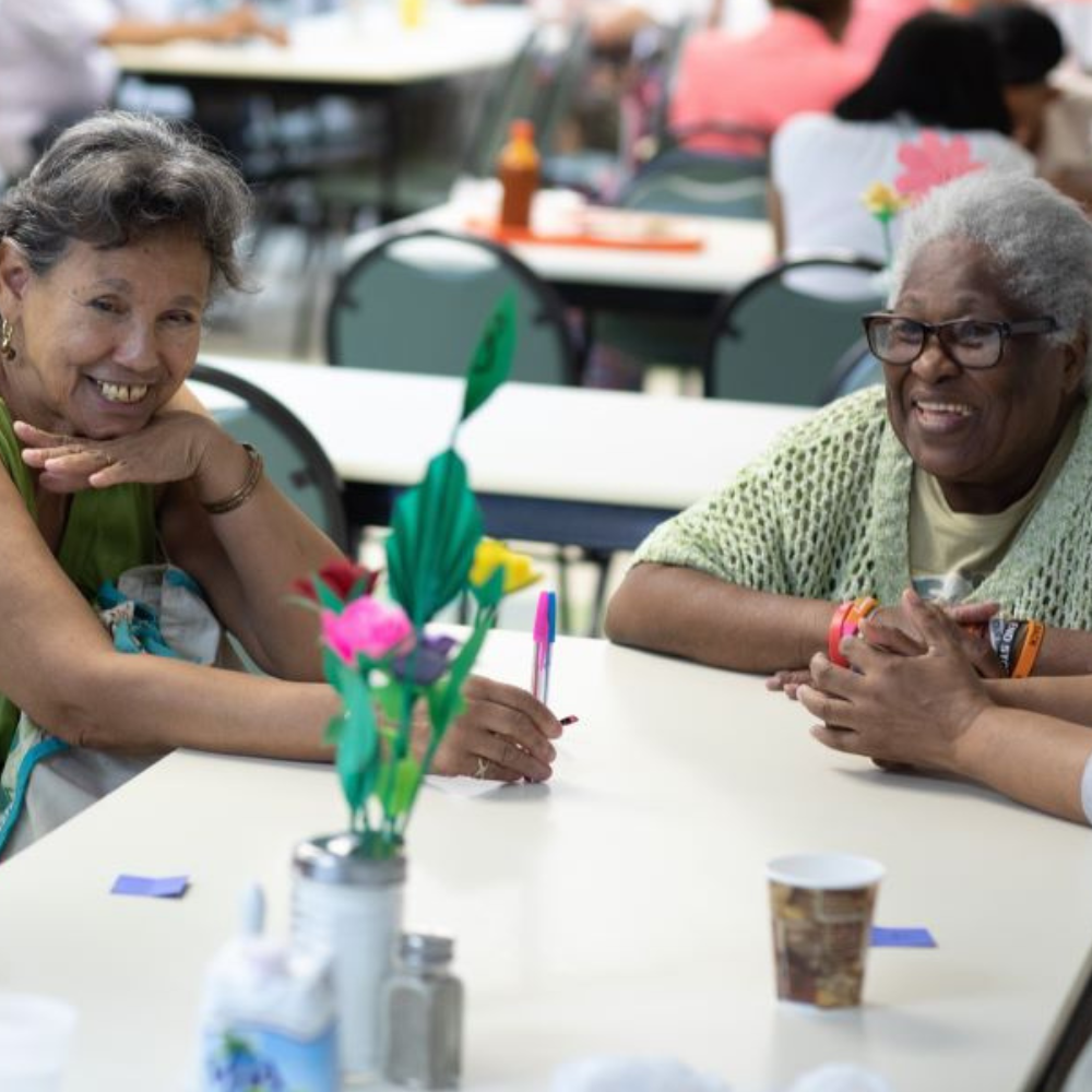 Two older adult women sit at a table and smile together.