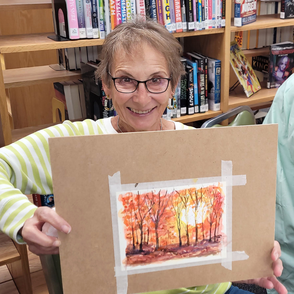 An older woman smiles to the camera and holds up a landscape painting.