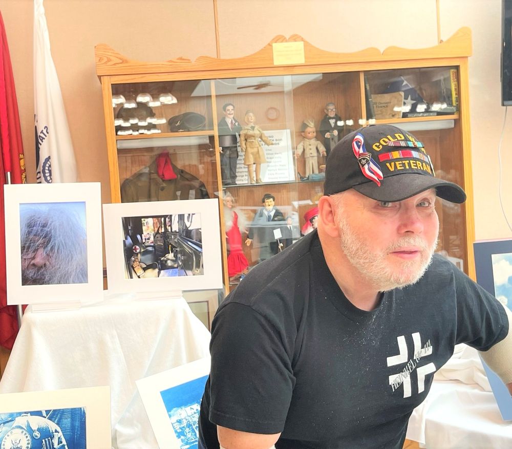 Older veteran man looks at the camera and poses with artwork. He is wearing a Cold War Veteran cap.