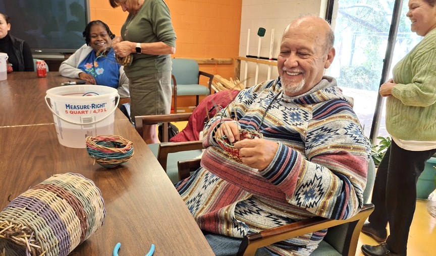 An older man smiles and sits in a chair while making a basket.