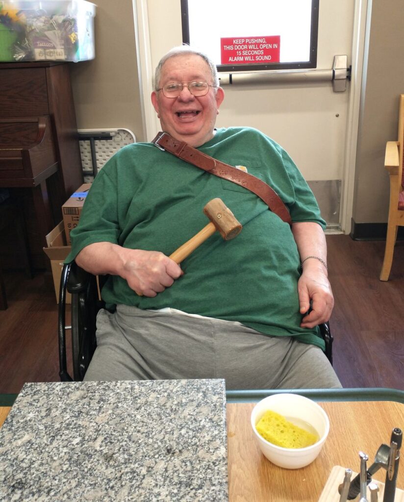 Older veteran man smiles joyfully at camera while holding a mallet. He has art materials in front of him on a table.