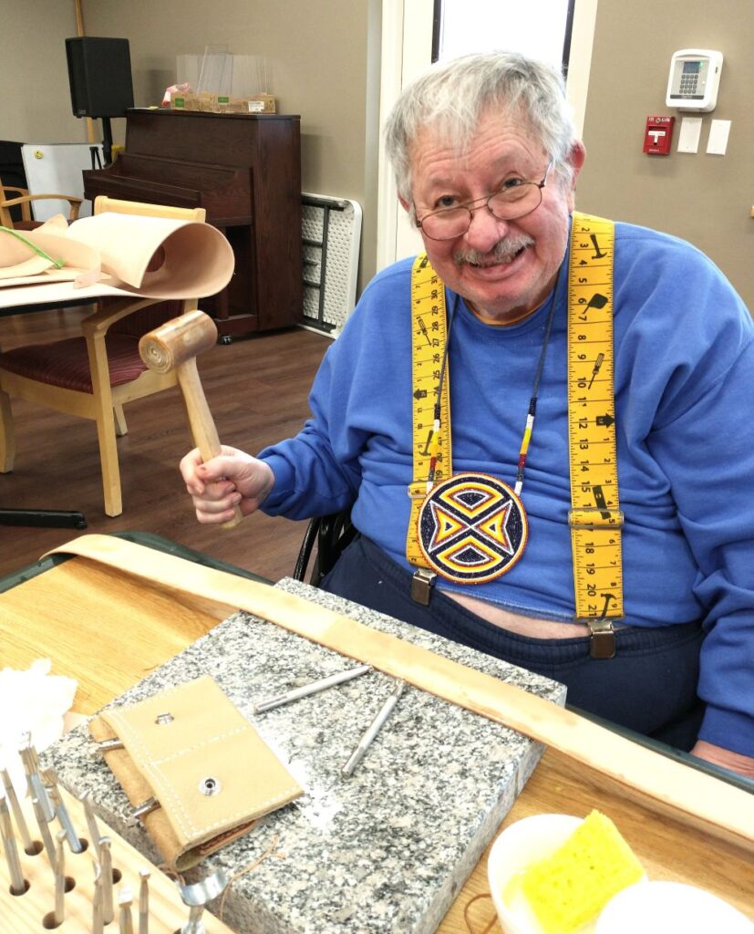 Older veteran man smiles with a mallet in hand. A concrete slate and leather belt is on the table in front of him.