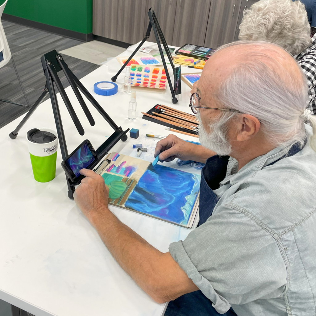 Older man paints at a table with a small table-top easel. 