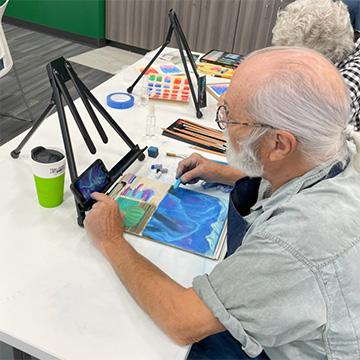 Older man paints at a table with a small table-top easel.