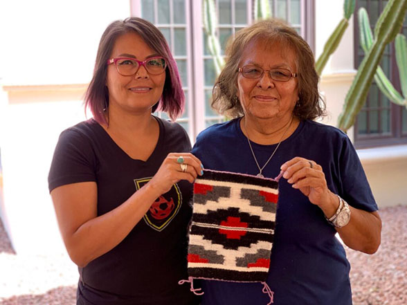 An older woman and younger woman stand next to eachother smiling while holding a small red, white, and black tapestry.