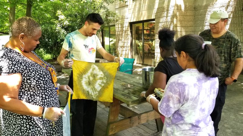 A younger man holds a natural dye cloth outside while children and older adults look at it.