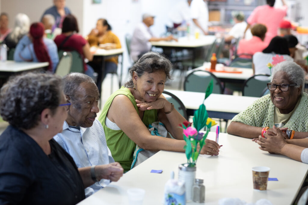 Four older adults of different racial backgrounds - 3 women and 1 man - enjoy each other's company around a table at a community center.