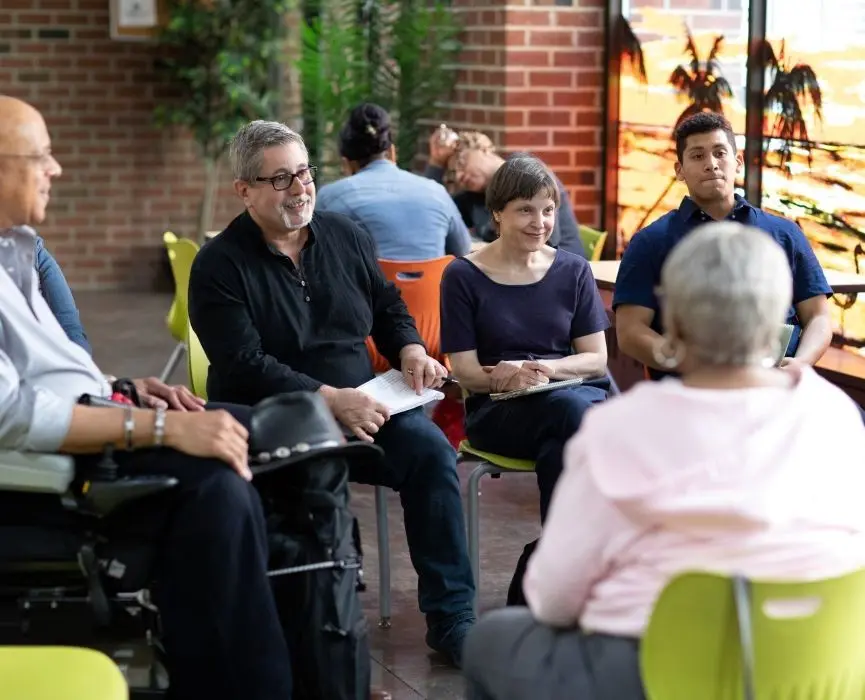 A group of older men and women sit in chairs in a circle smiling and talking.