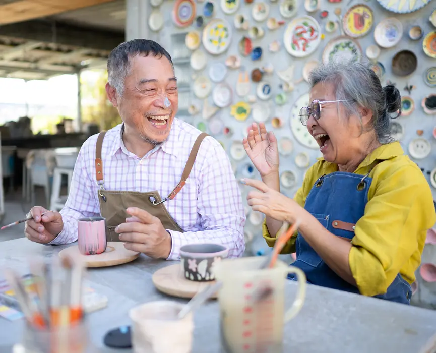An older man and woman sit at a table working on pottery. They're both laughing and smiling at each other.