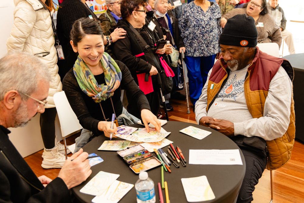 A Black older adult man, white older adult man, and Asian middle-aged woman work on an art project at a circular table together. The Asian woman and Black older adult man are smiling.