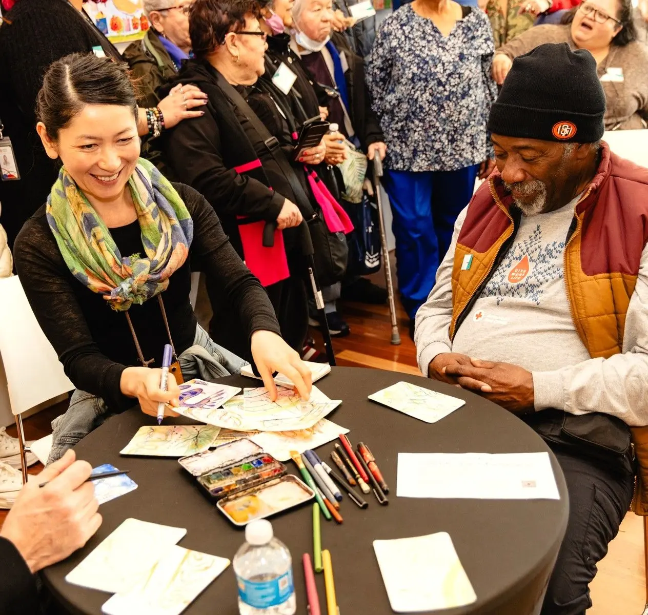 A younger woman and older man sit together at a round table while making art.