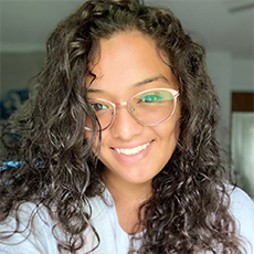 Headshot of Daniela Dale Velez. She has long, curly brown hair. She is smiling and wearing gold-framed glasses.