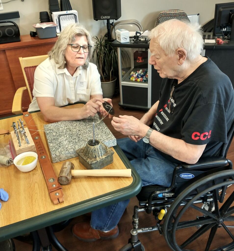 Older veteran man works on a leather belt at a table with a woman instructor. The instructor is showing the veteran a tool.