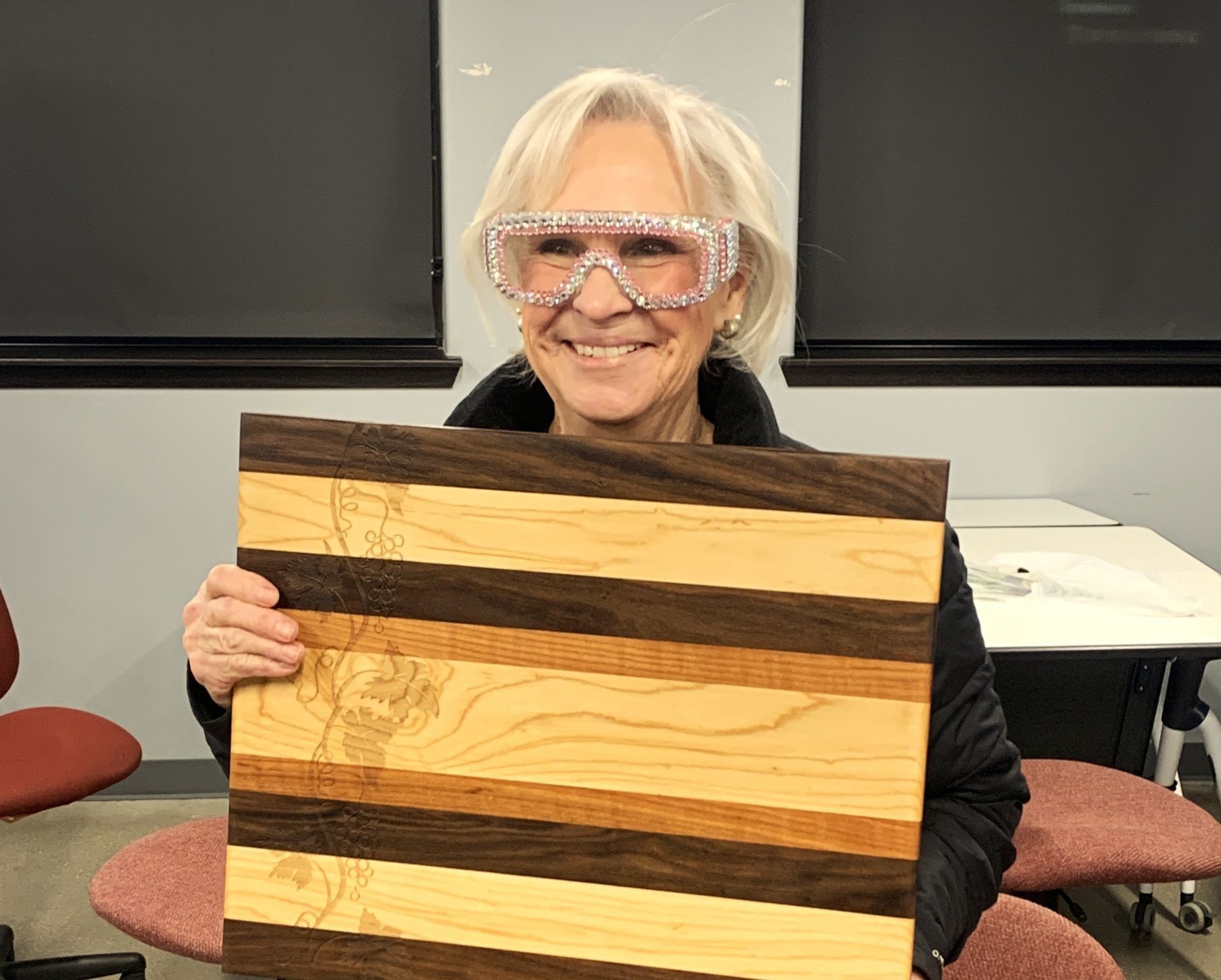Dixie, a creative aging program participant, holds up her carved woodworking project and smiles with goggles on.