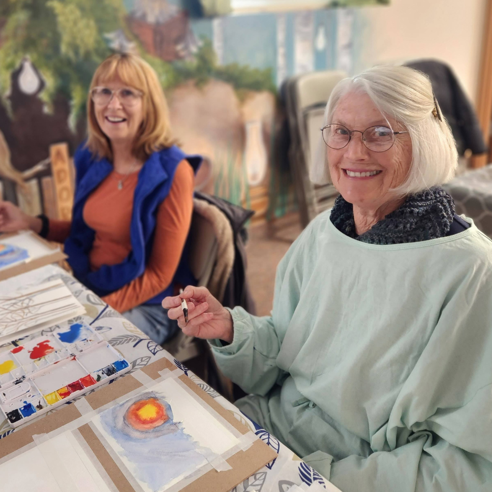 Two older woman smile while painting at a table.