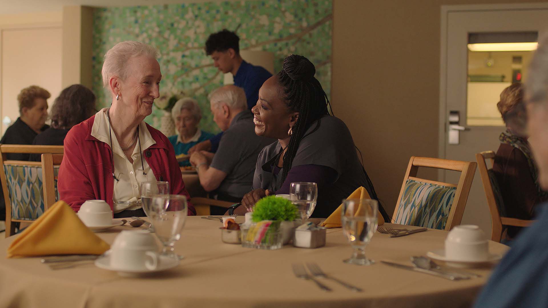 An older adult woman talks to a younger female caregiver at a dining table.