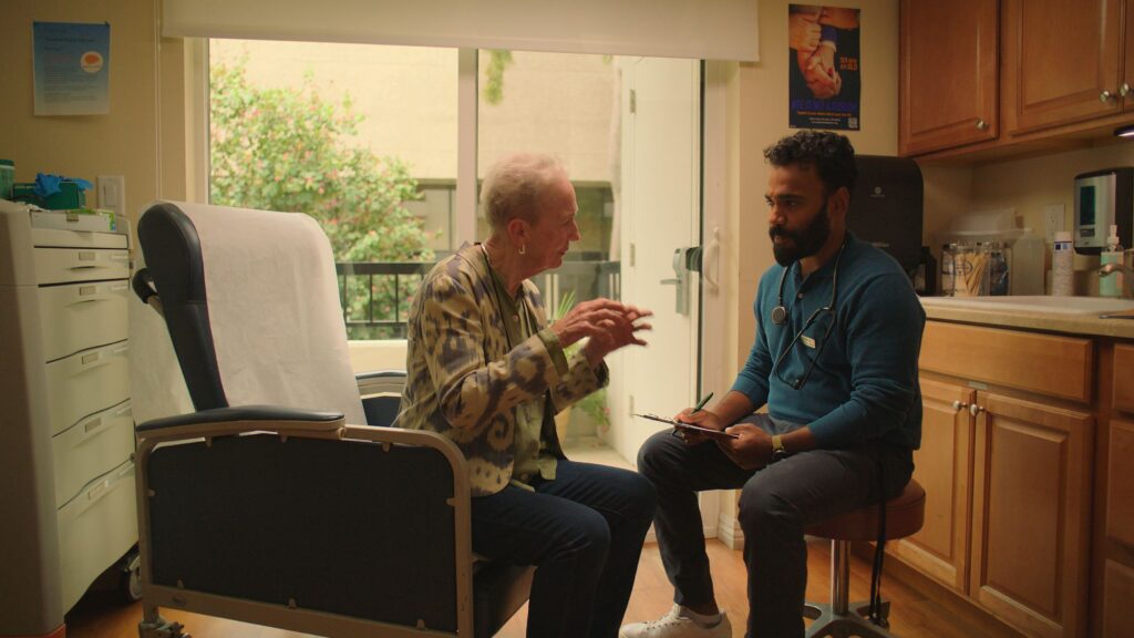 A still from the film, "Familiar Touch." It shows an older woman white short, white hair talking with her hands to a younger man whose a caregiver. The man has dark curly hair and a beard.