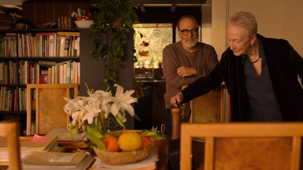 A still from the film, "Familiar Touch." It shows an older woman picking something up on a table and a younger man behind her staring at her.