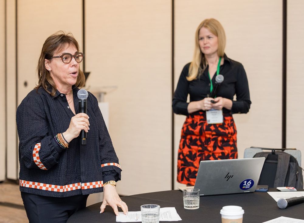 A middle-aged woman speaks with a microphone. Heather Ikemire, Lifetime Arts' Executive Director, stands in the background listening.