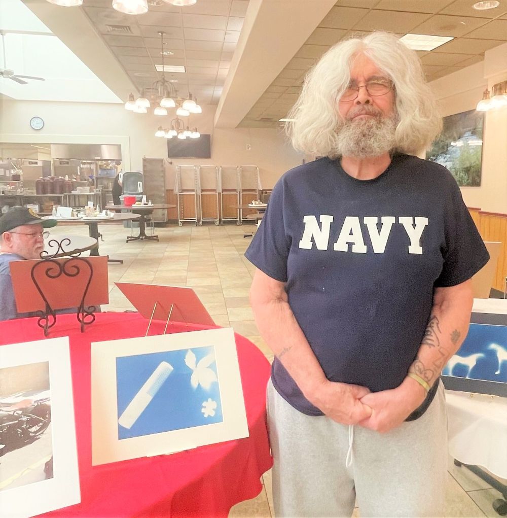 Older veteran man smiles and poses with his artwork. He is wearing a Navy t-shirt.