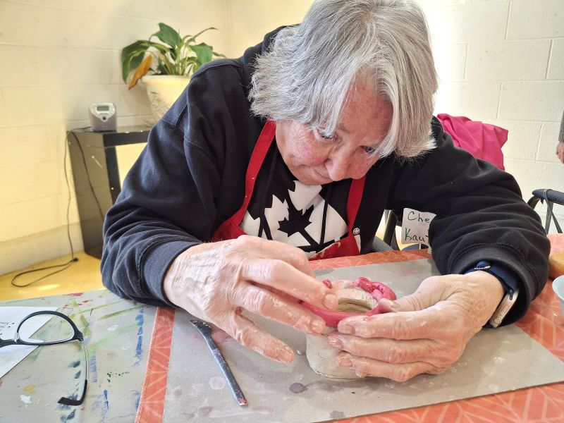 An older woman sits in a chair and intently works on a piece of clay.