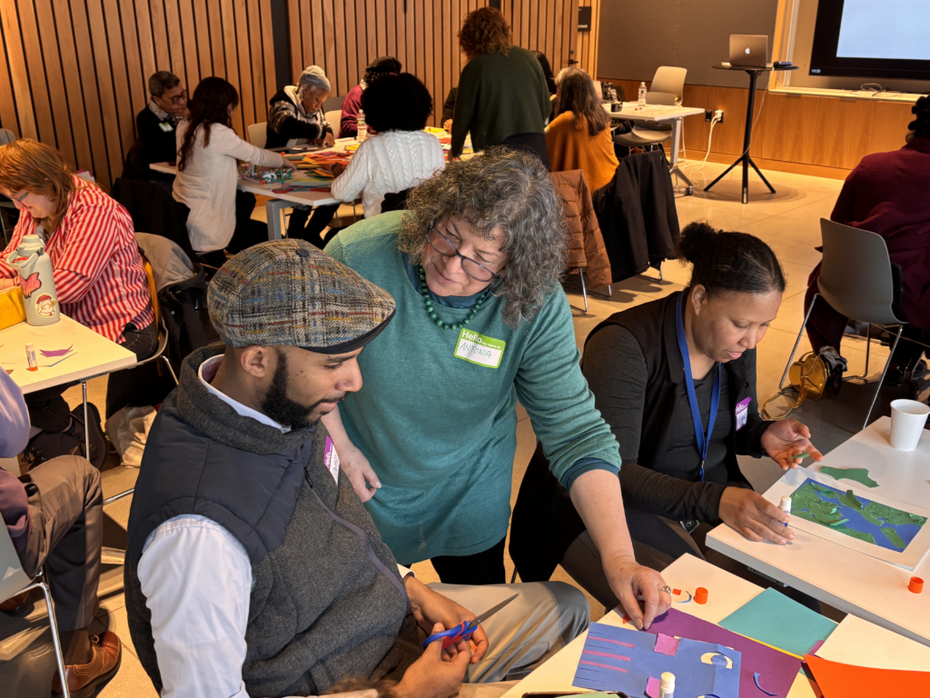 A teaching artist stands next to a participant sitting at a table, looking at his collage together.