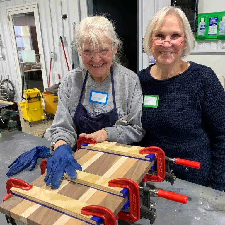 Two older women pose next to each other while one has their hand on a piece of woodwork. 