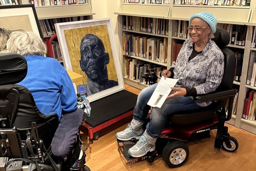 A Black older adult woman smiles and holds a piece of paper in front of a wall of books. She is in a wheelchair.