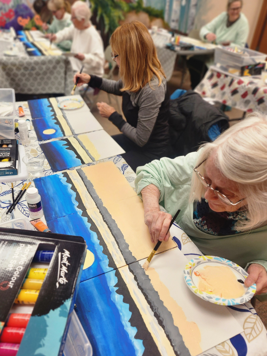 Two older woman paint a landscape at a table.