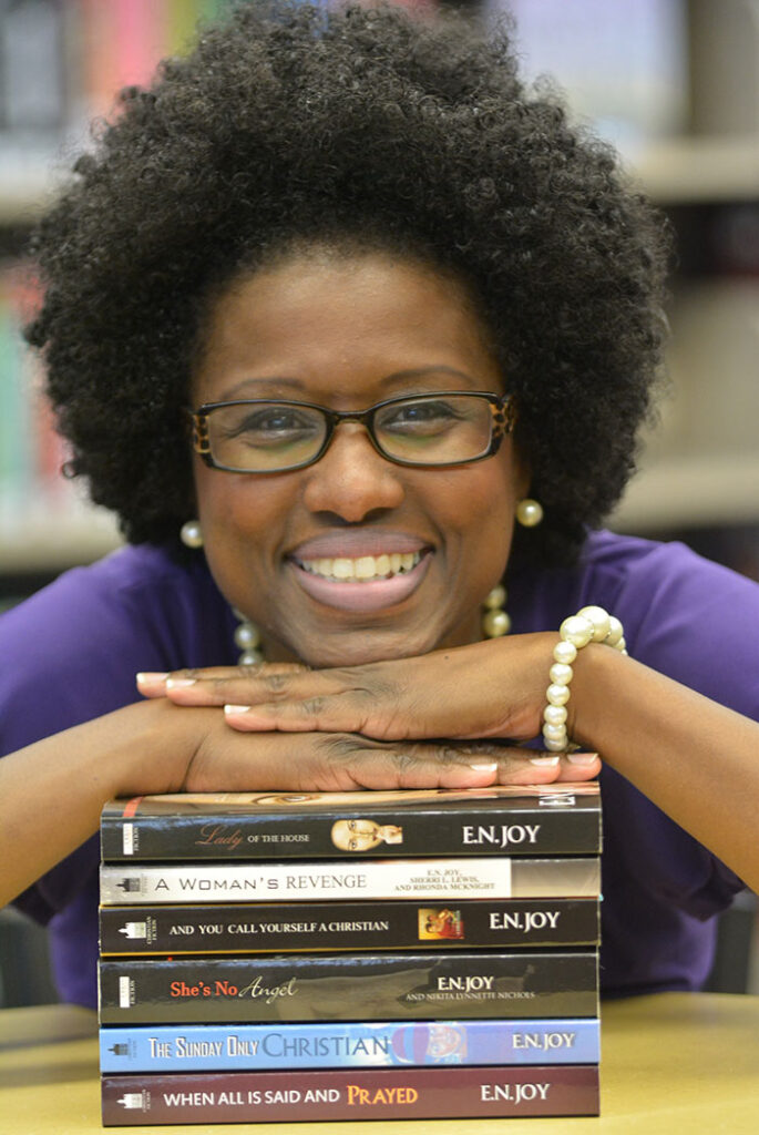 Headshot of Joylynn M. Ross. She has dark curly hair and dark framed glasses. She is resting her chin and hands on a stack of books on a table.