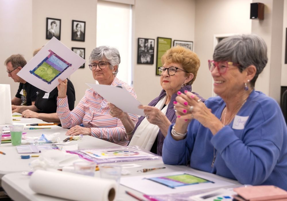 Three older adult women sit at a table together with painting in front of them. They smile while one of them holds up her painting.