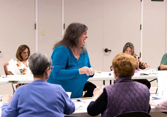 A teaching artist is surrounded by students at tables. She laughs and looks off camera.
