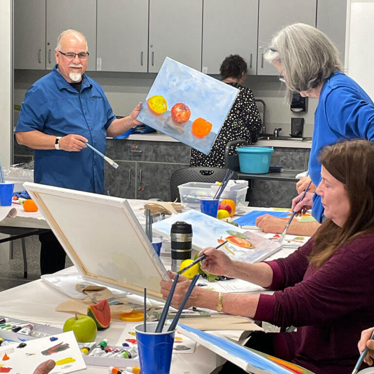An older man holds a painting while looking at an older woman, whose back is to the camera.