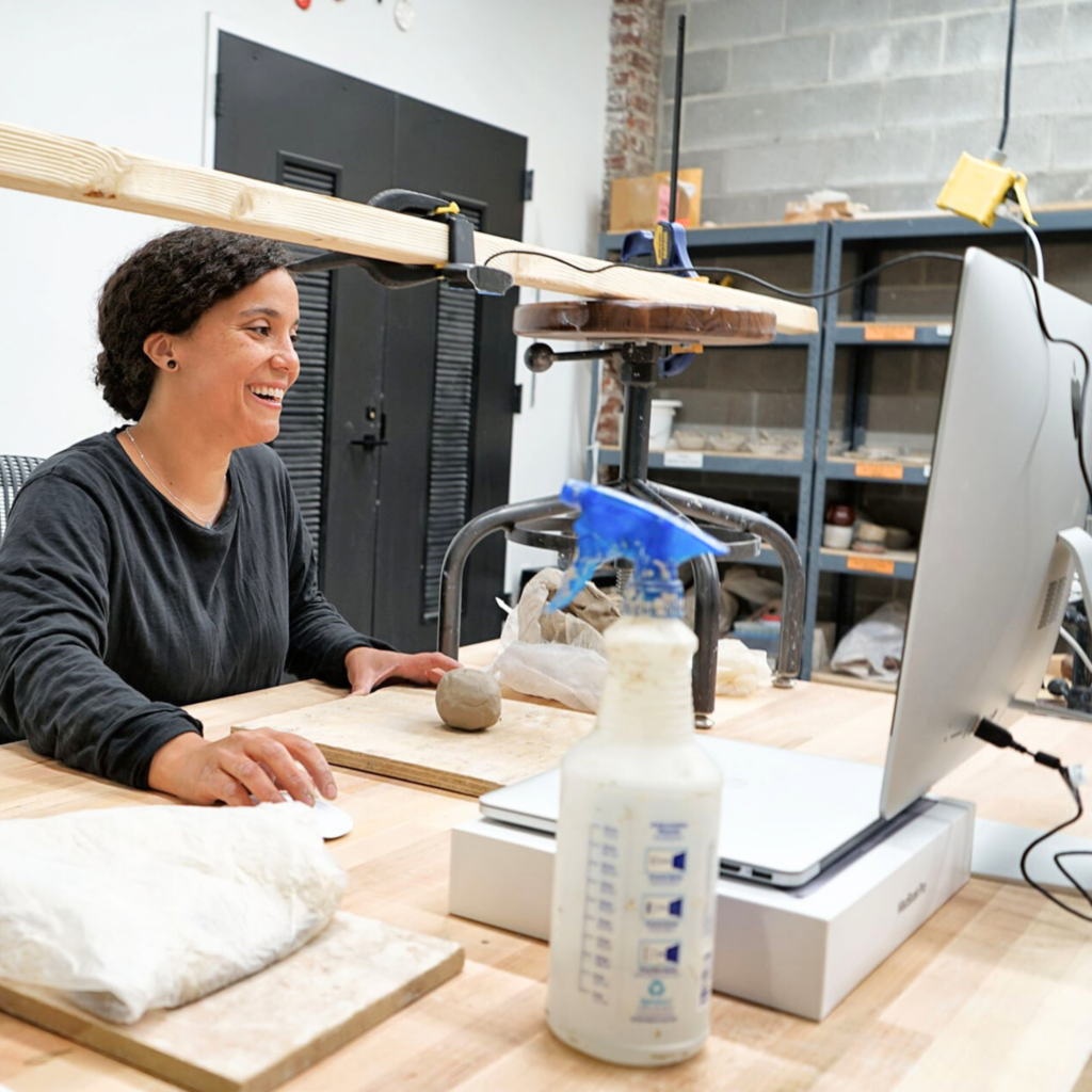 A younger woman smiles while using a computer. A ball of clay is placed in front of her.