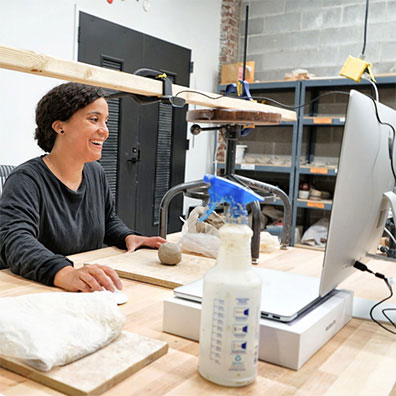 A younger woman smiles while using a computer. A ball of clay is placed in front of her.