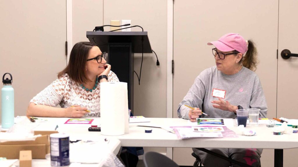 Liza Cucco, Director of Strategic Communications, sits at a table next to an older adult student during a creative aging program. They are both looking at each other and smiling.