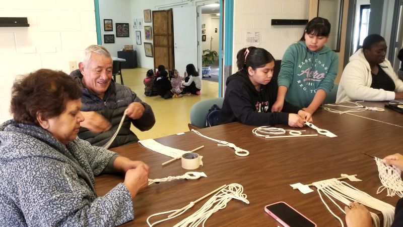 A group of older adults and children work on macrame with mustard seed pieces. 