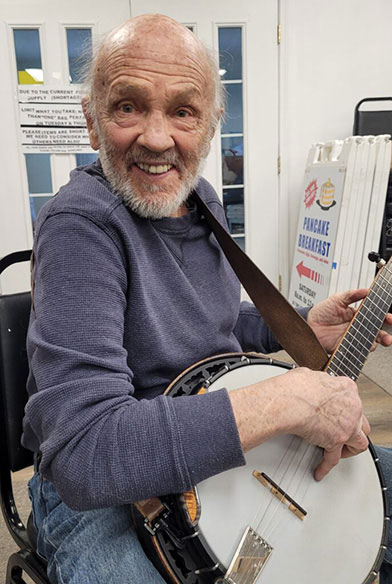 An older adult man plays a guitar and smiles at the camera.